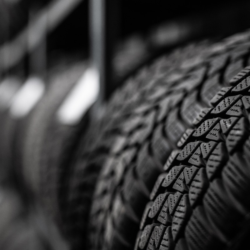 Gros plan monochrome sur la bande de roulement texturée d'un pneu de voiture, avec d'autres pneus flous en arrière-plan.