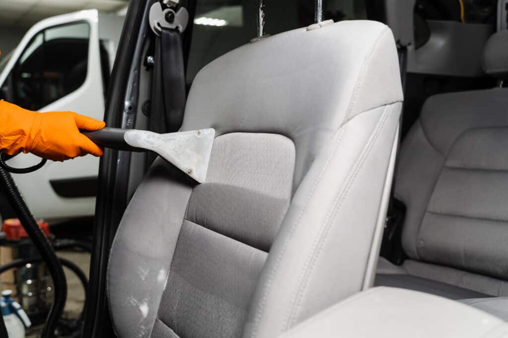 Nettoyage Siège Auto : Intérieur Voiture Impeccable Main gantée de orange nettoyant un siège auto gris avec un extracteur, des zones mouillées apparentes. Nettoyage intérieur professionnel.