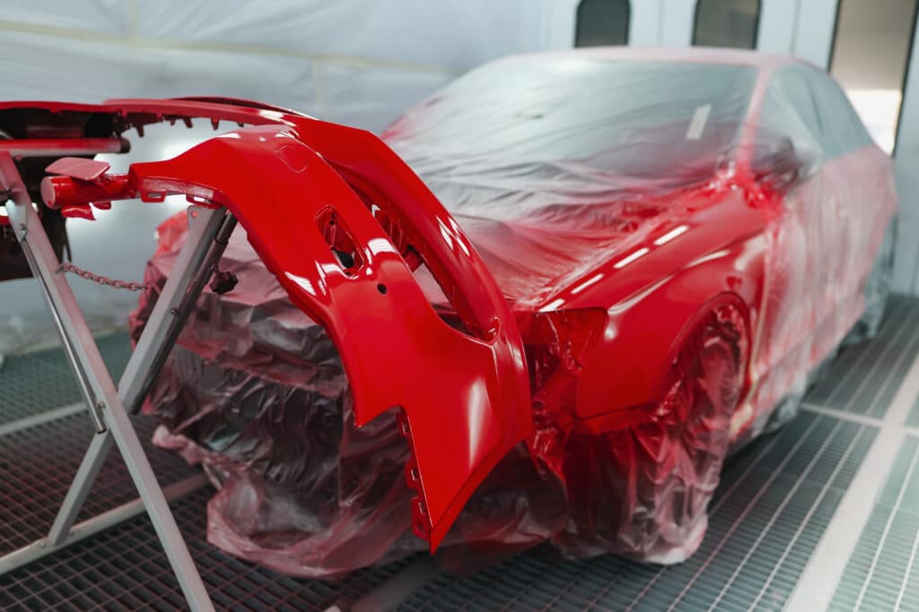 Peinture voiture rouge : carrosserie et pare-chocs éclatants Voiture rouge et pare-chocs fraîchement peints en rouge vif, séchant dans une cabine de peinture automobile. La carrosserie est protégée par une bâche plastique.