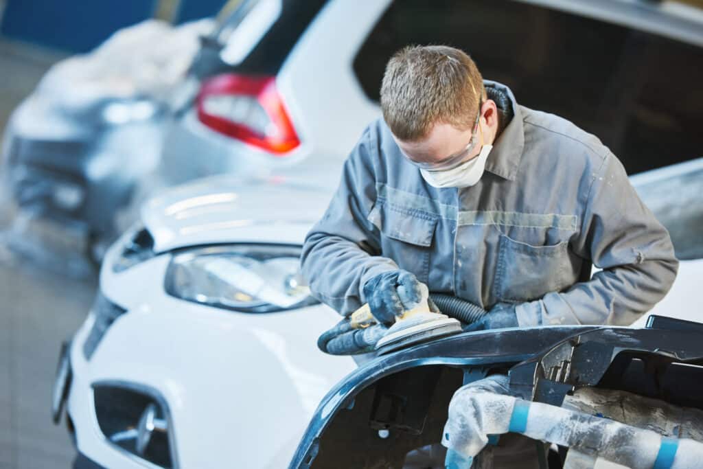 Un homme ponce un pare-chocs de voiture blanc avec une ponceuse orbitale. Il porte un masque, des lunettes et des gants pour la sécurité.