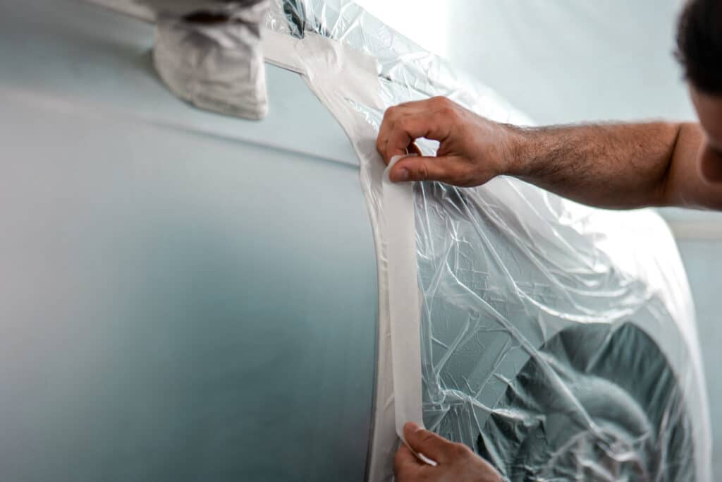 Préparation peinture voiture : masquage de vitre avec ruban adhésif Mains appliquant du ruban de masquage sur une bâche plastique transparente recouvrant la vitre d'une voiture gris clair avant peinture.