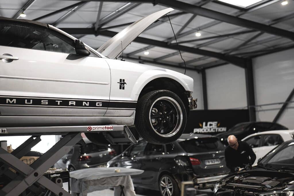 Réparation Ford Mustang en atelier | Entretien Automobile Mustang blanche sur pont élévateur Primetool, capot ouvert. Mécanicien travaillant sur moteur. Garage LCE Production.