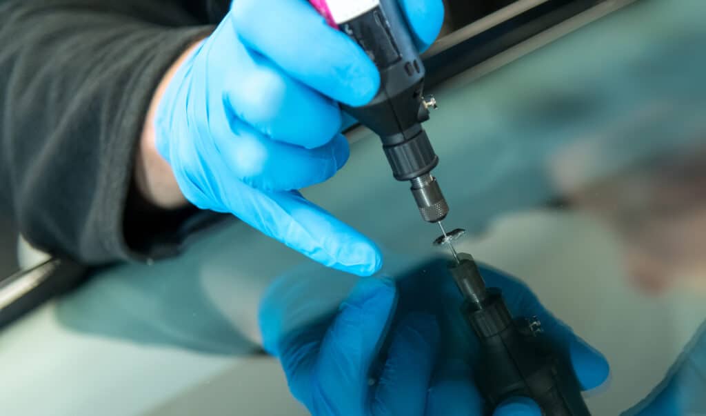 Mains gantées de bleu réparant un impact sur un pare-brise. Un outil injecte de la résine dans le verre, avec un reflet visible.