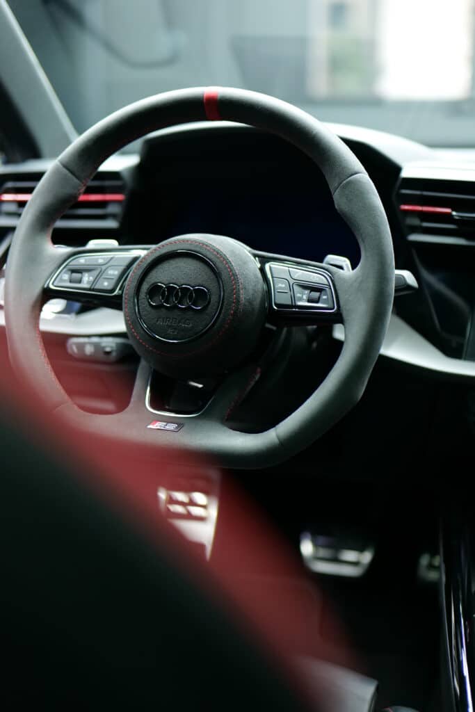 Volant Audi RS Sport en Alcantara : Performance et Luxe Volant sport Audi RS en Alcantara noir avec surpiqûres rouges, bande rouge à 12h et logo Audi. Commandes multifonctions et "AIRBAG" visibles.